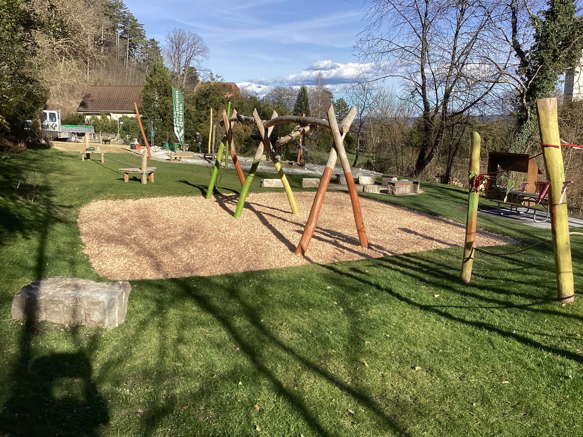 Ein kleiner, terrassenförmig angelegter Wasserfall mit aufgeschichteten Felsen, üppigen tropischen Pflanzen und einer Holzterrasse in einem sonnigen Garten.