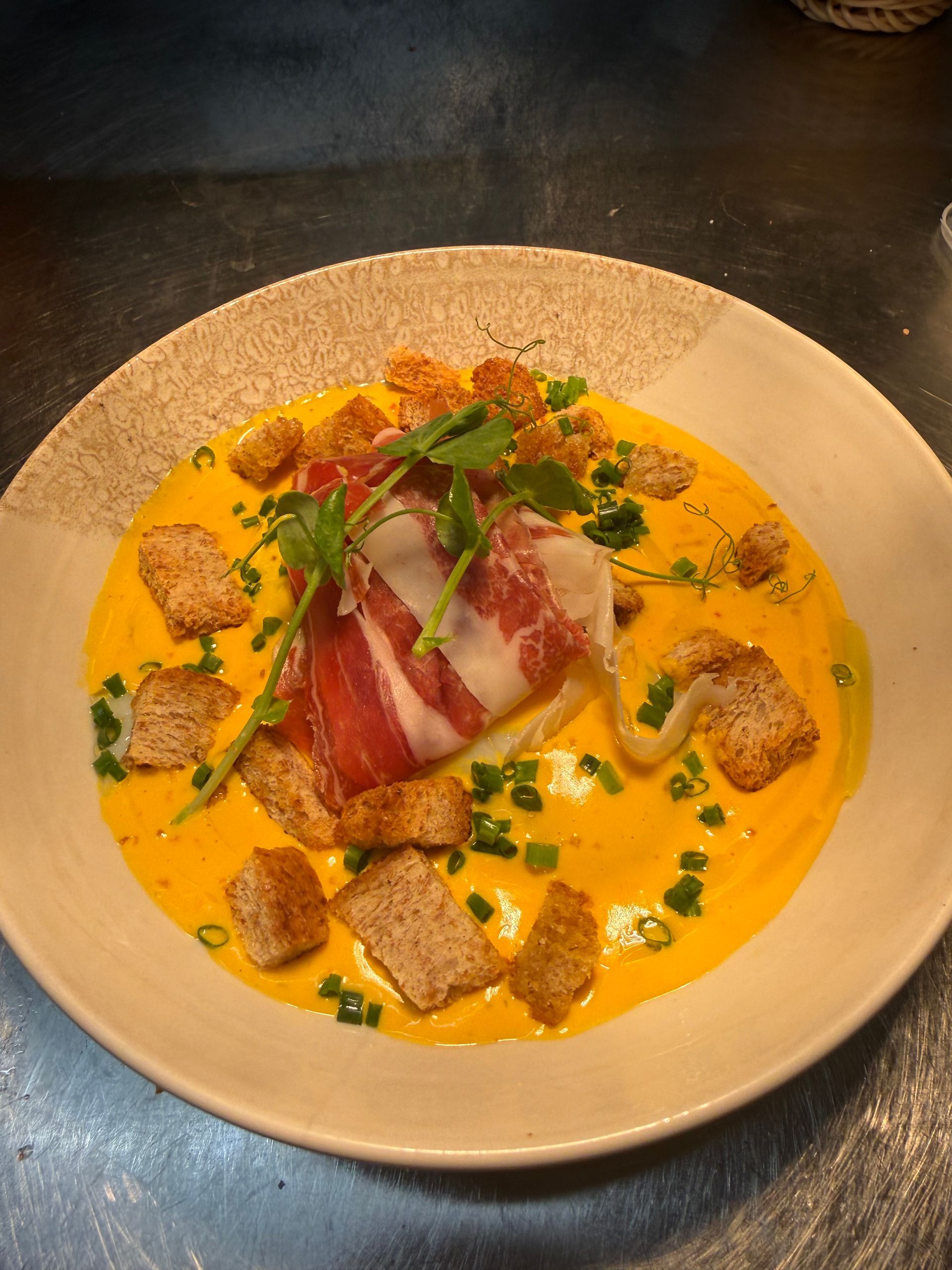 Soupe crémeuse à l'orange dans un bol, garnie de prosciutto, de croûtons et d'herbes.