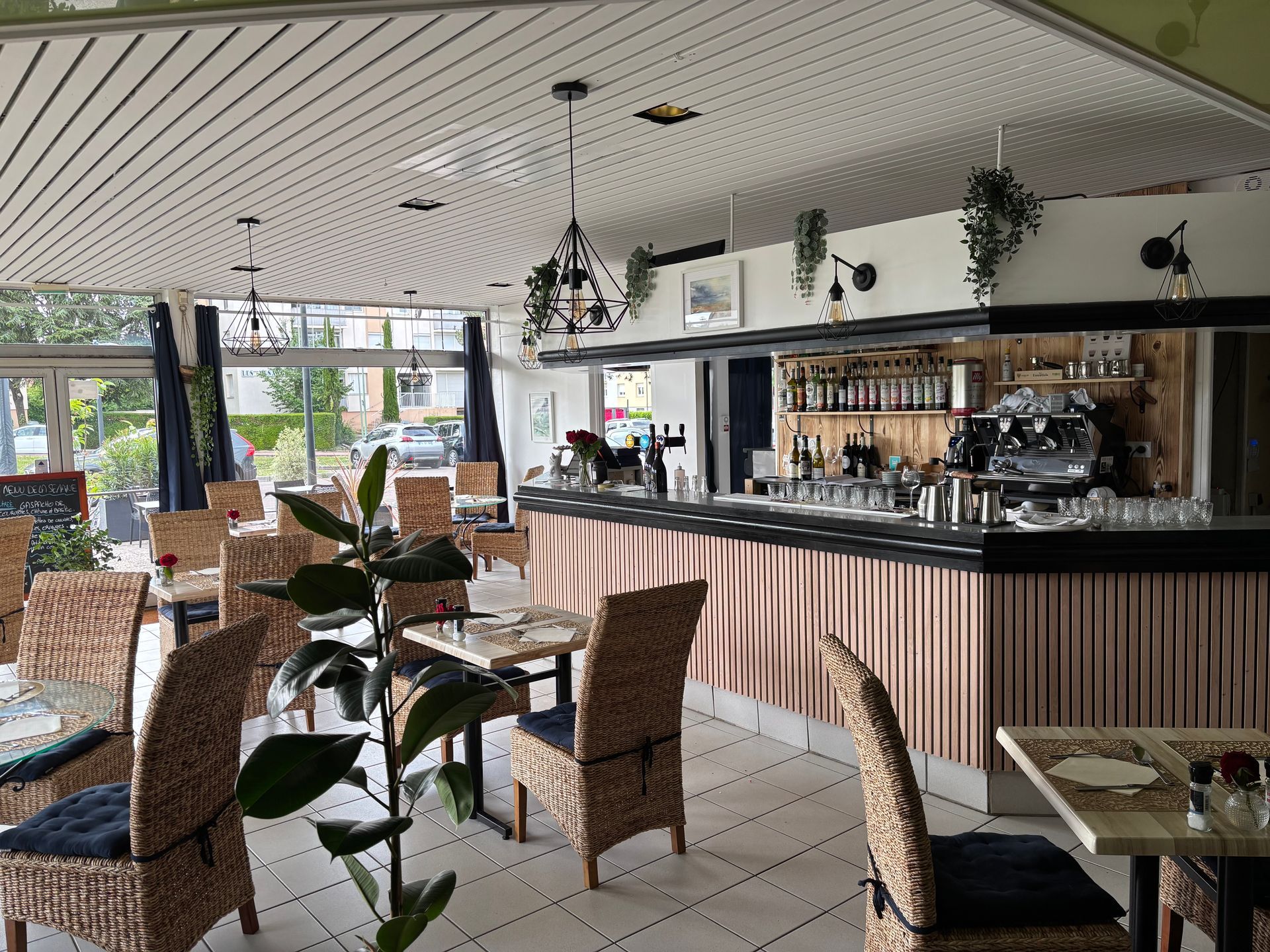 Vue intérieure du restaurant L'ENVIE avec des chaises en osier, un bar et des plantes, avec une palette de couleurs neutres.