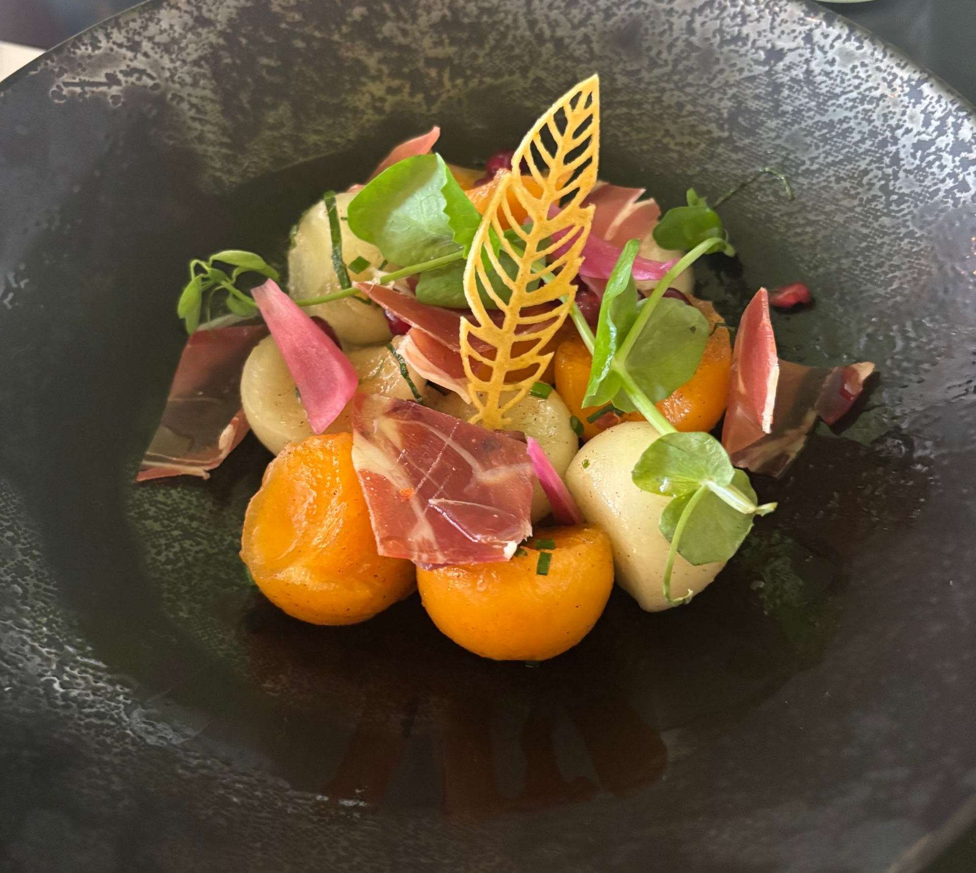 Une salade colorée avec des boules de melon, du jambon et une feuille décorative sur une assiette sombre.