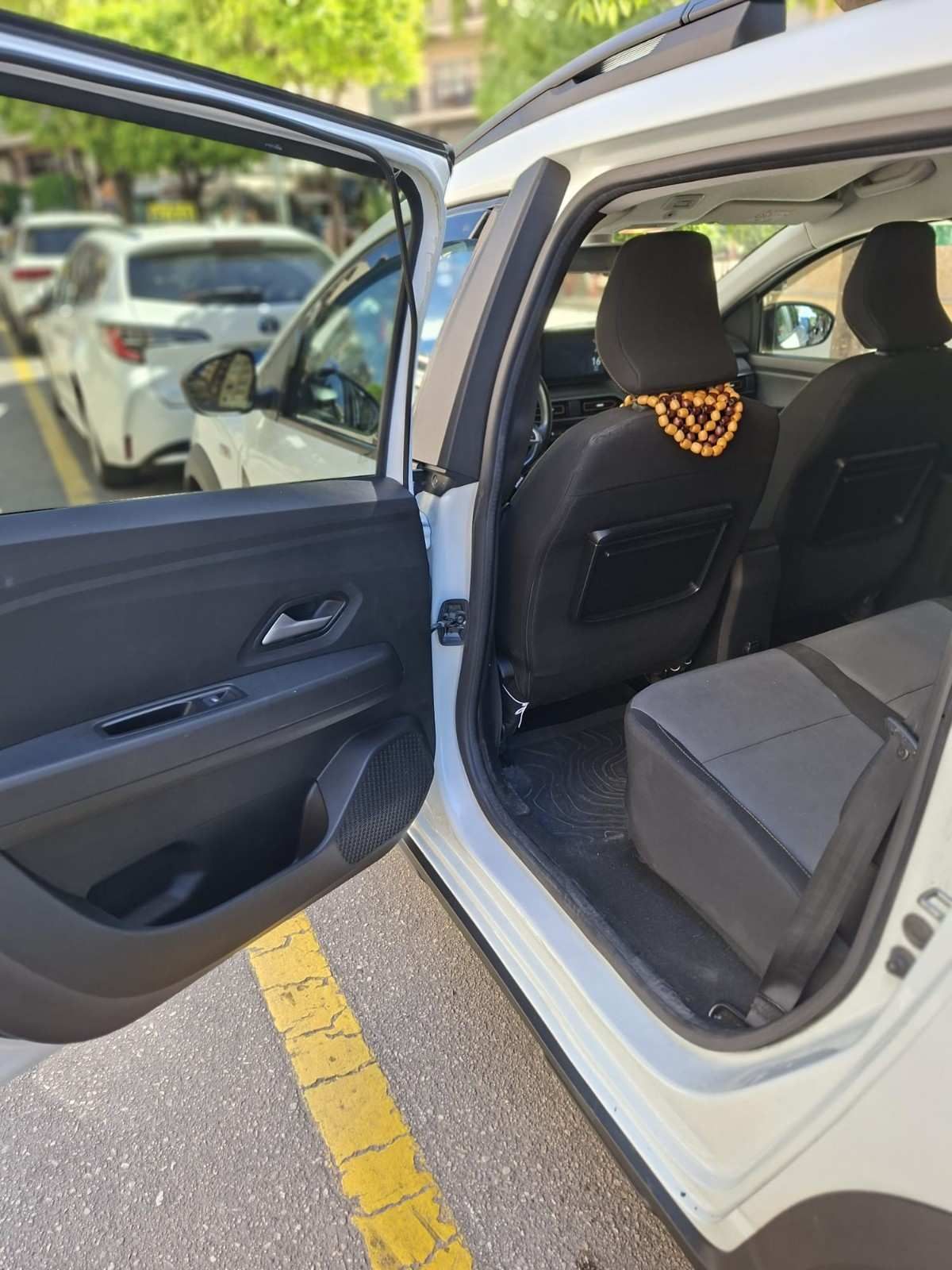 La puerta trasera abierta de un coche blanco, que deja ver los asientos interiores grises, un reposacabezas negro y una cubierta con cuentas.