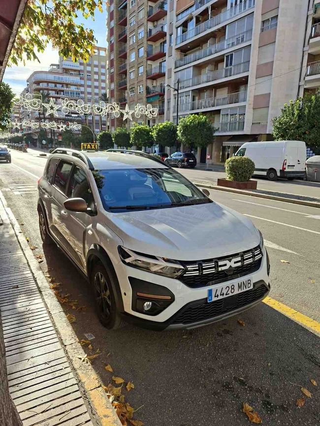 Un SUV Dacia Jogger blanco estaba estacionado al costado de una calle arbolada de la ciudad, junto a edificios de apartamentos.
