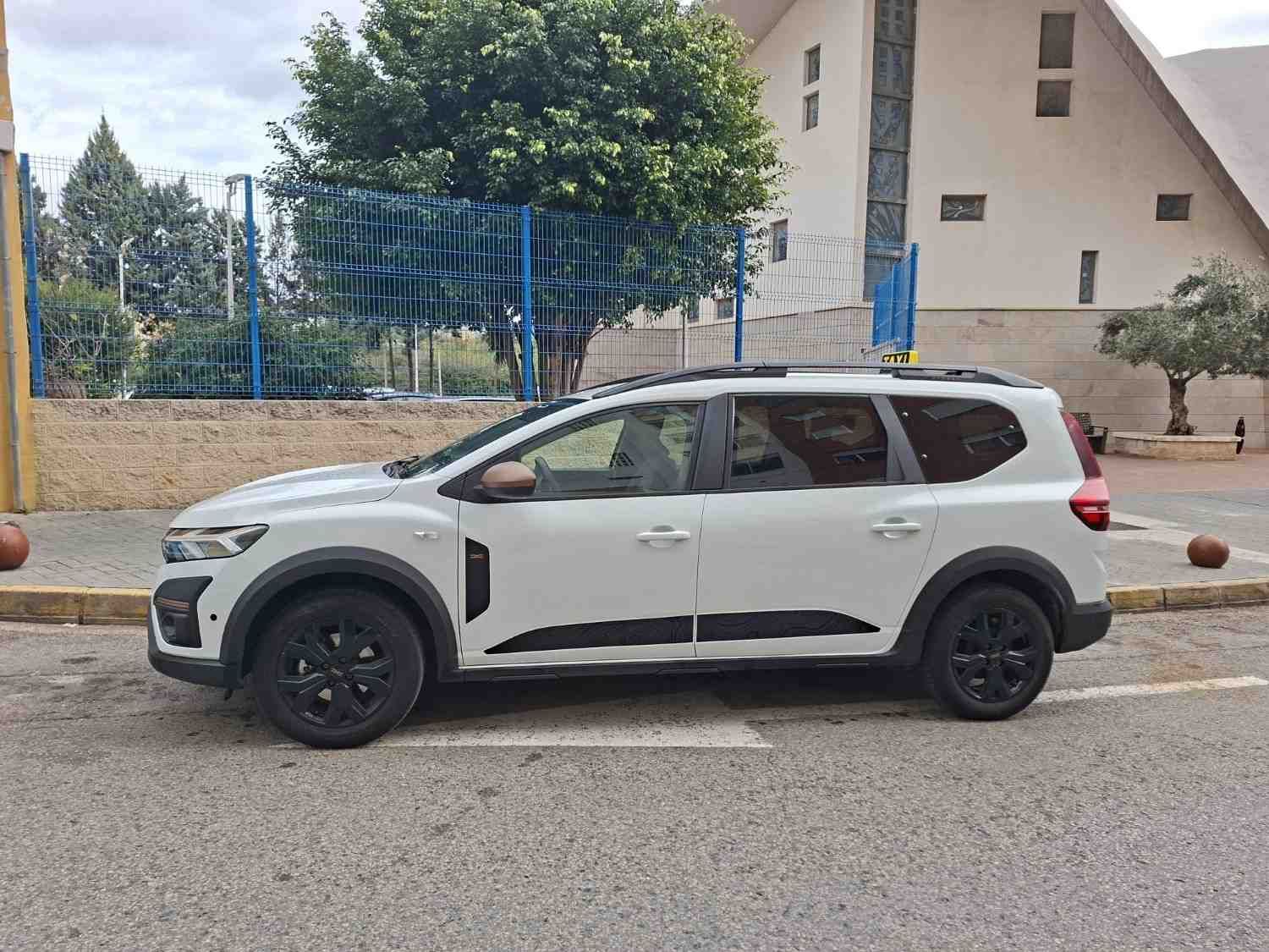Un SUV Renault Triber blanco estacionado en un terreno pavimentado junto a un edificio y unos árboles.