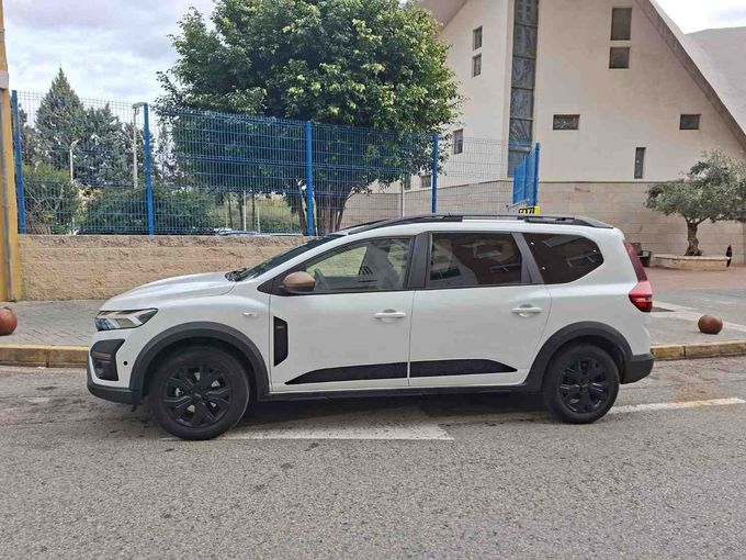 Un SUV Renault Triber blanco estacionado en un terreno pavimentado junto a un edificio y unos árboles.