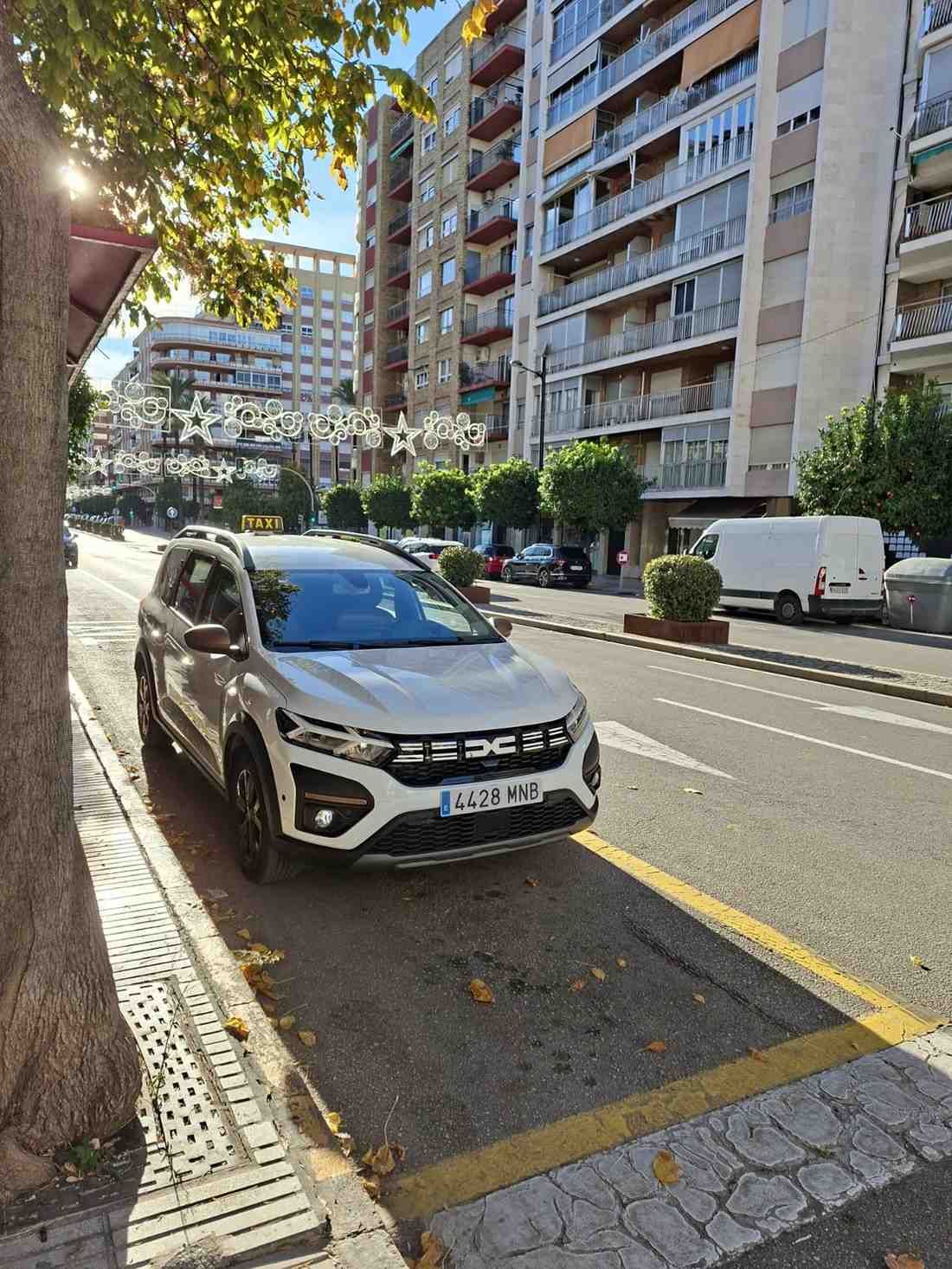 Un Dacia Jogger blanco aparcado al lado de una calle urbana bordeada de árboles y edificios de apartamentos.