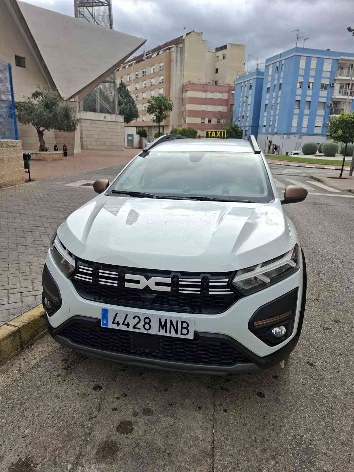 Un taxi Dacia Sandero Stepway blanco estaba estacionado en una calle pavimentada frente a un edificio con un gran tejado triangular.