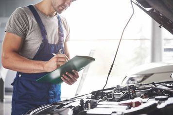 Un hombre mira debajo del capó de un automóvil mientras sostiene un portapapeles.