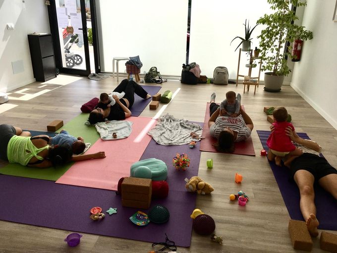 Clase de yoga con adultos y bebés en una sala llena de luz;