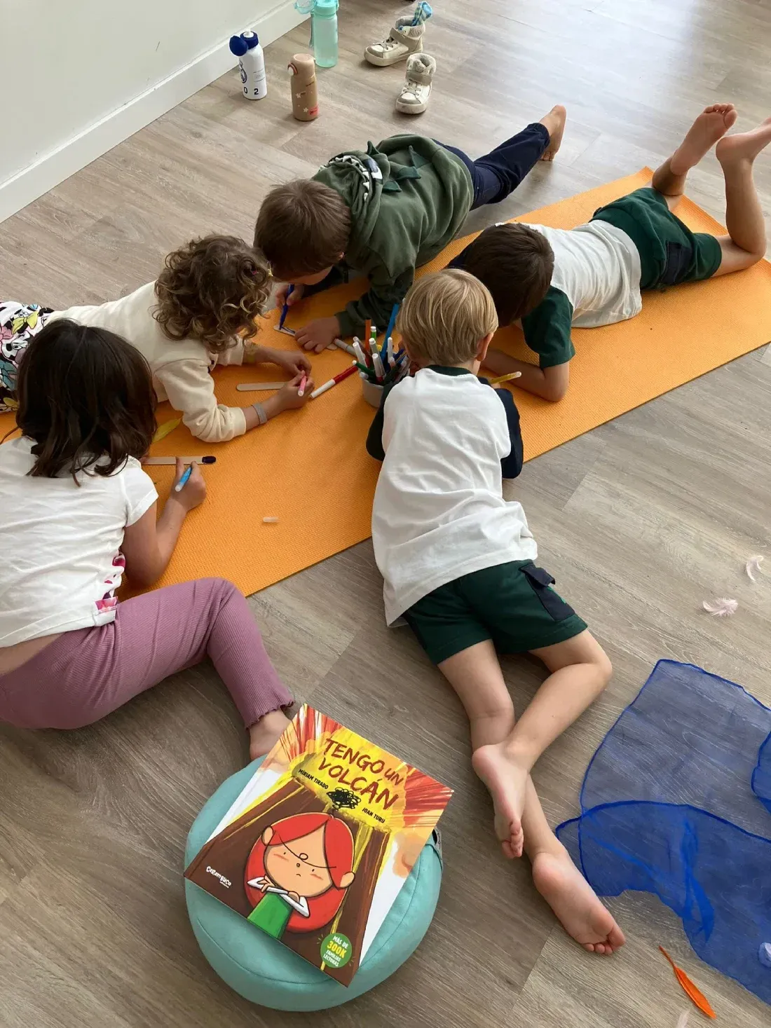 Niños dibujando sobre un tapete naranja, tumbados en el suelo. Uno lee un libro, mientras otros dibujan con rotuladores.