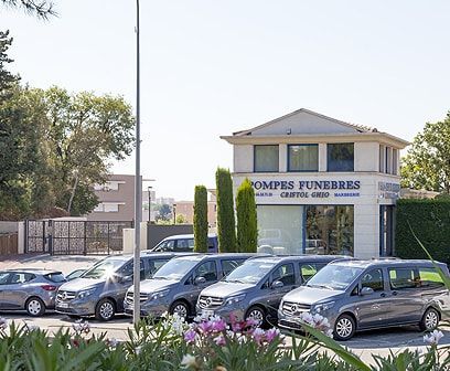 Bâtiment funéraire avec une flotte de fourgons gris garés devant. Enseigne bleue, journée ensoleillée.