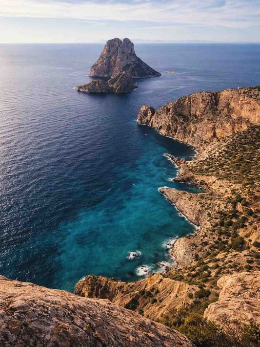 Acantilados rocosos con vistas a una cala turquesa y una isla lejana bajo un cielo despejado.