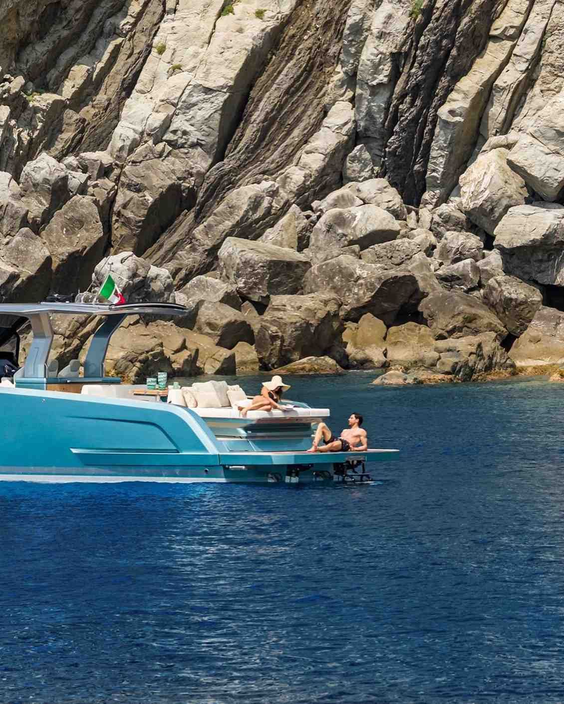 Un yate turquesa con gente nadando a su lado cerca de acantilados rocosos junto al mar.
