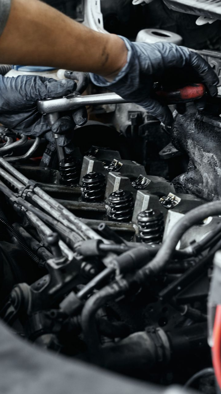 Mécanicien travaillant sur un moteur de voiture, portant des gants. Compartiment moteur avec pièces, outils et câbles sombres.