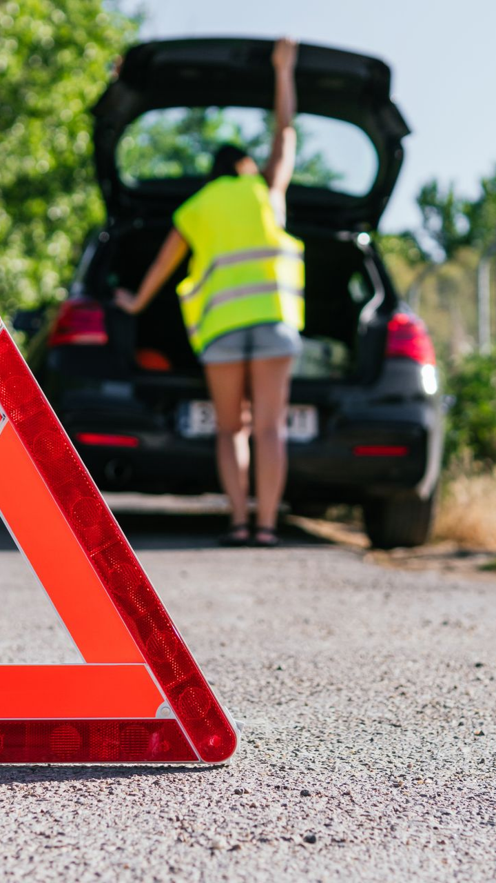 Femme portant un gilet réfléchissant près d'une voiture, coffre ouvert, au bord de la route. Triangle d'avertissement rouge au premier plan.