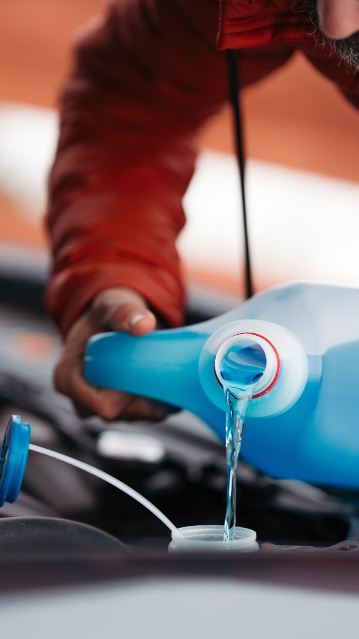 Une personne verse du liquide lave-glace bleu dans le réservoir d'une voiture, veste rouge visible.