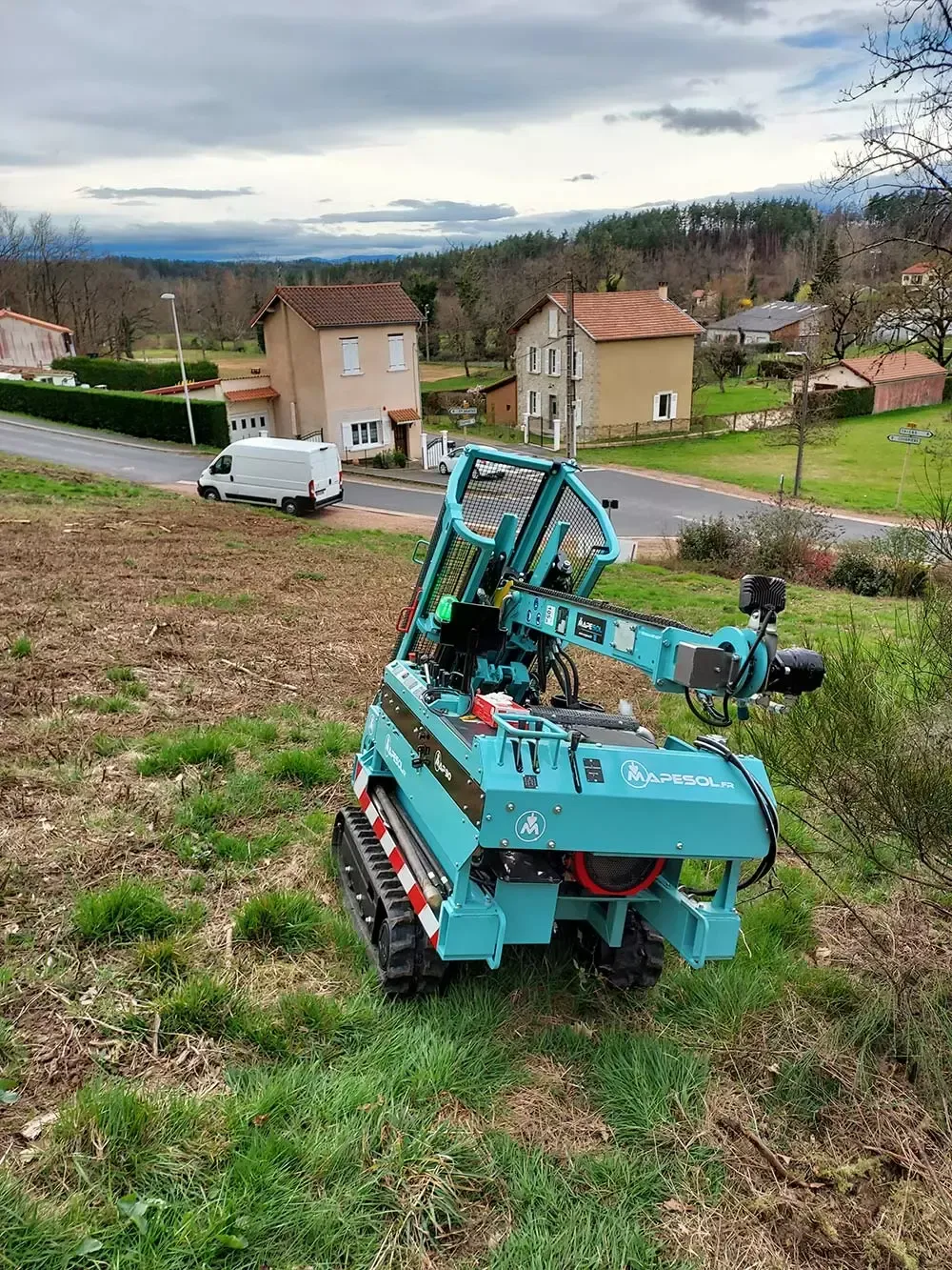 Une machine forestière robotisée de couleur sarcelle sur des chenilles dans un champ herbeux, des maisons et des arbres en arrière-plan.