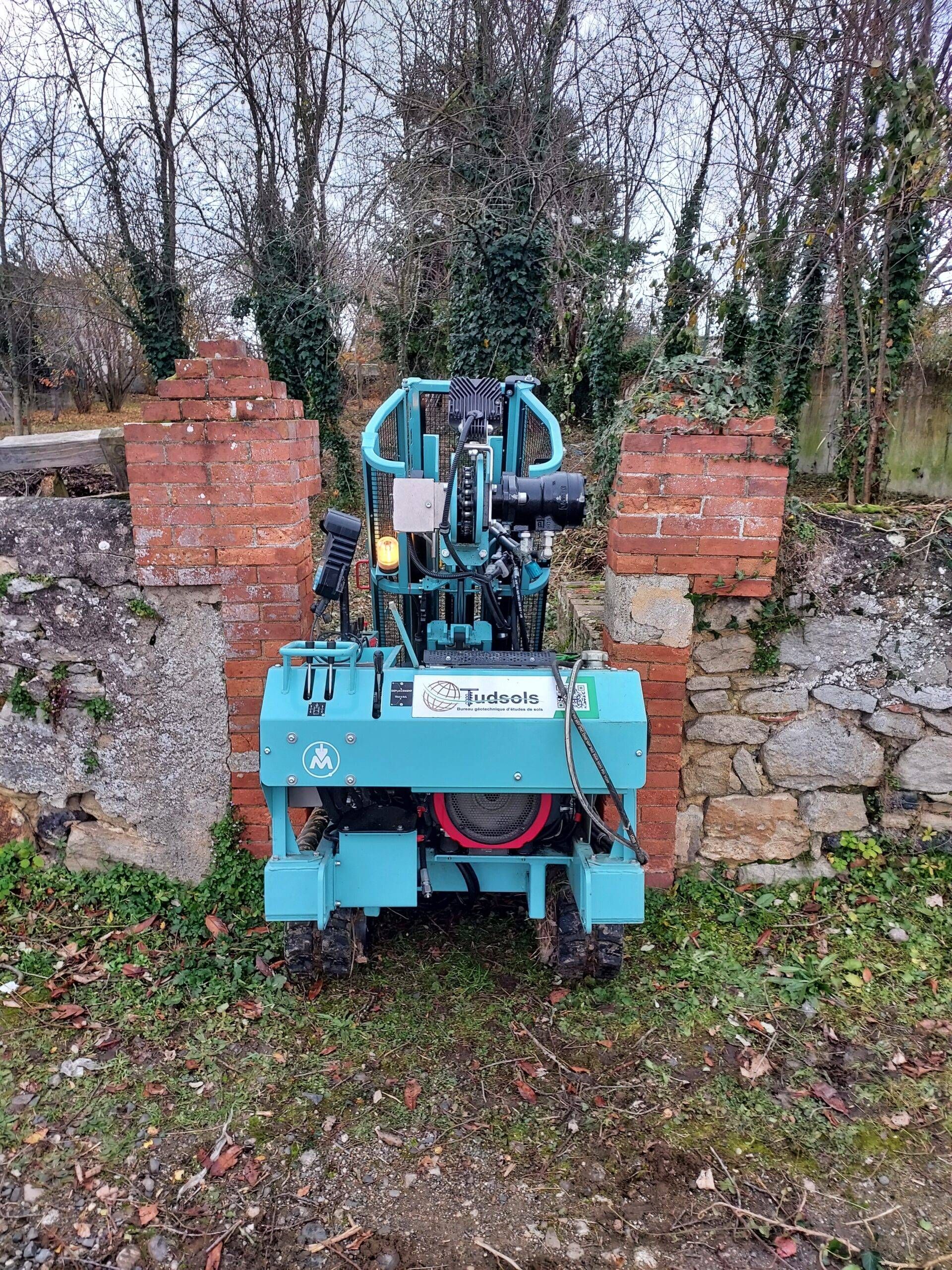Une machine de couleur sarcelle avec une roue rouge se trouve devant des ruines de briques et de pierre, de l'herbe verte au premier plan.