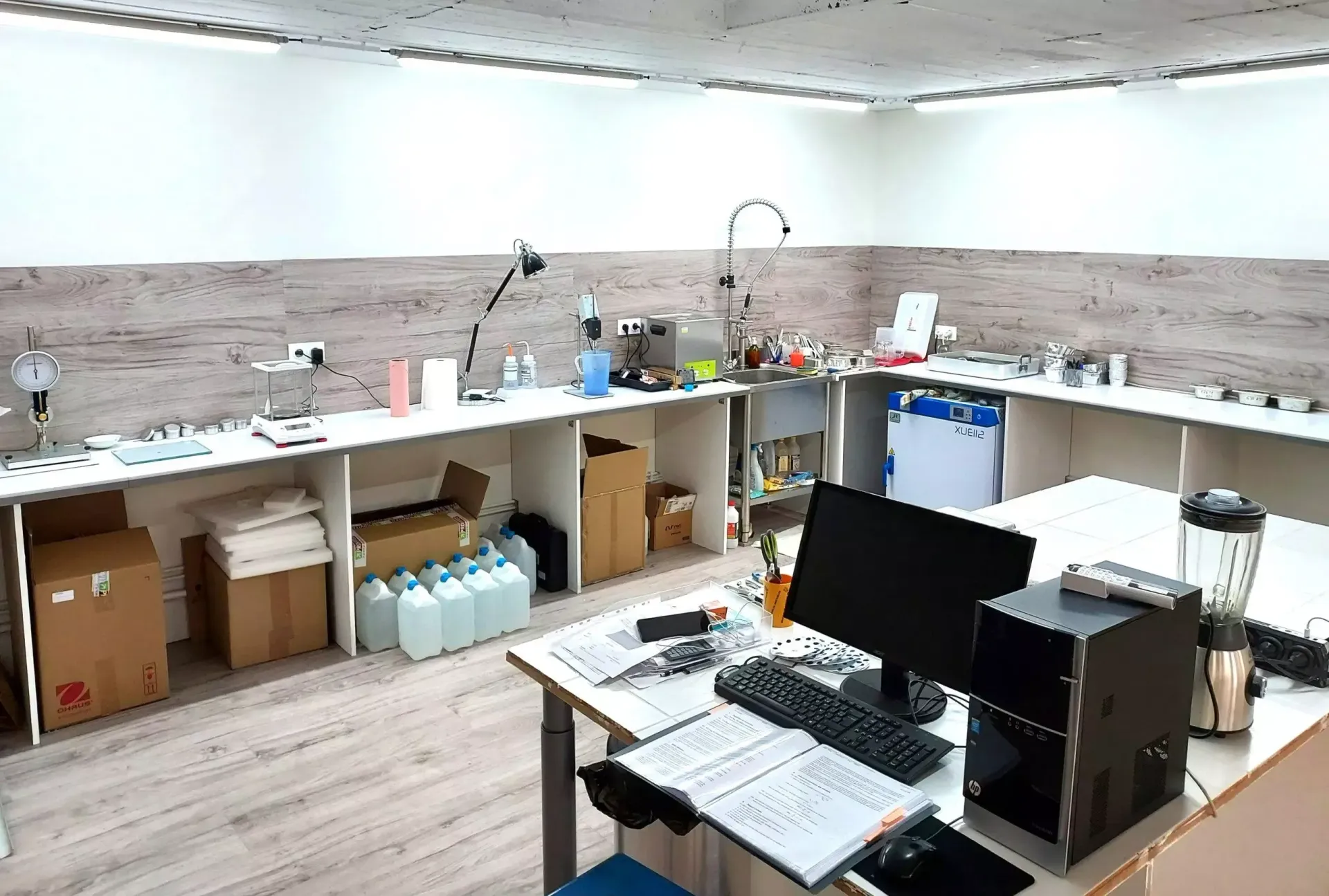 Laboratoire avec comptoirs blancs encombrés, ordinateur et divers équipements.