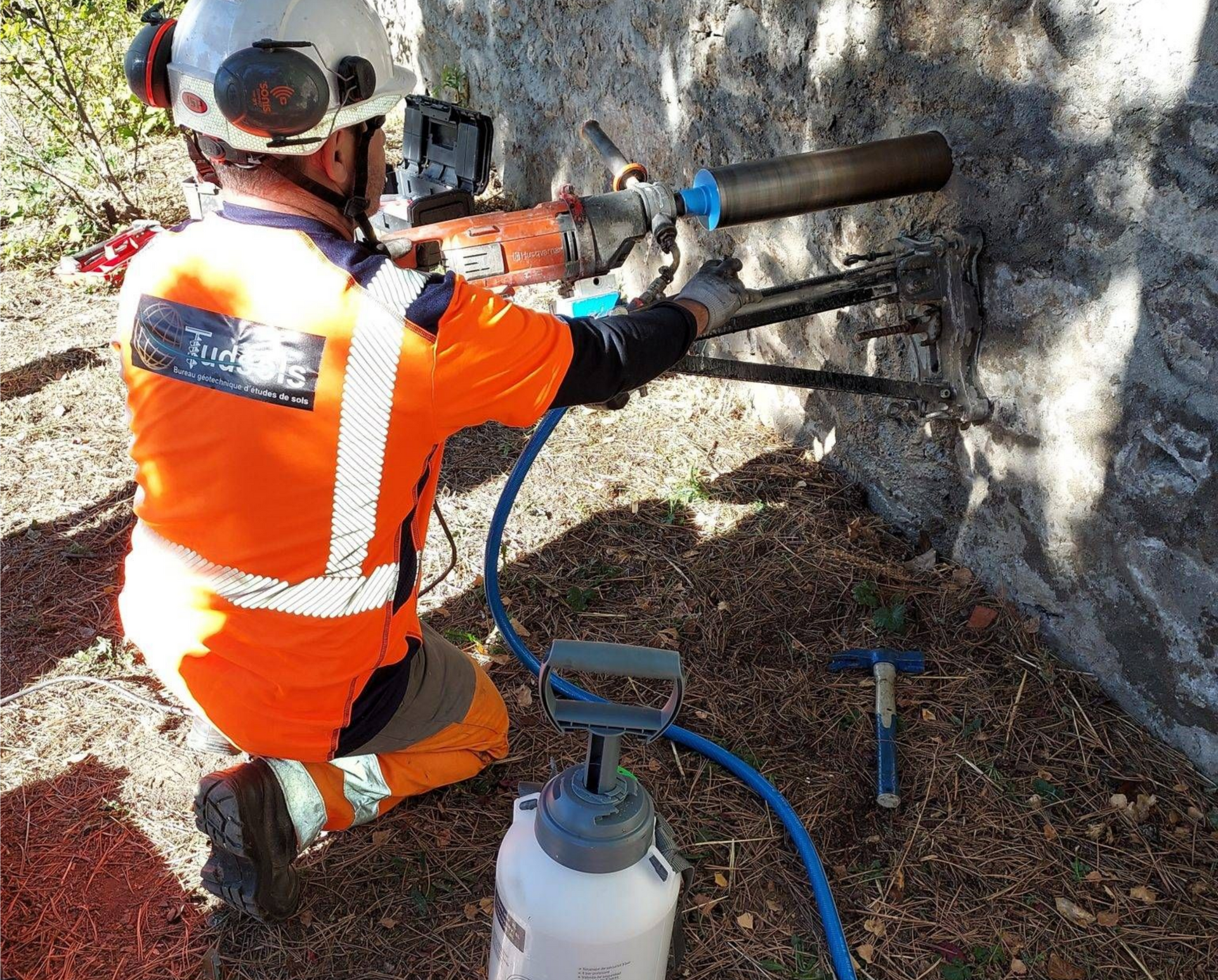 Un ouvrier perce un mur de pierre avec un équipement de sécurité et un gilet orange.