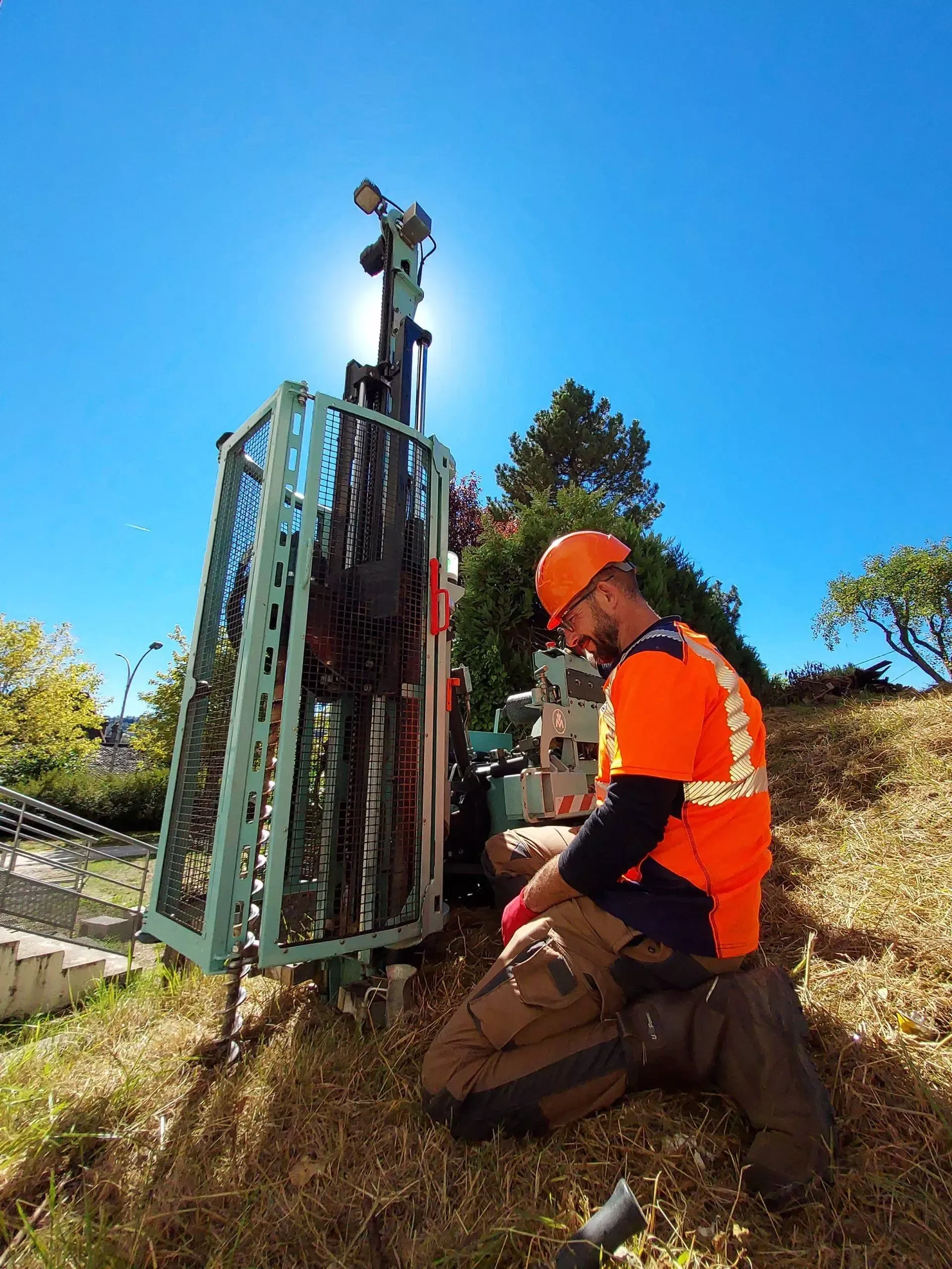 Un ouvrier en gilet orange s'agenouille près d'une perceuse sur une colline.