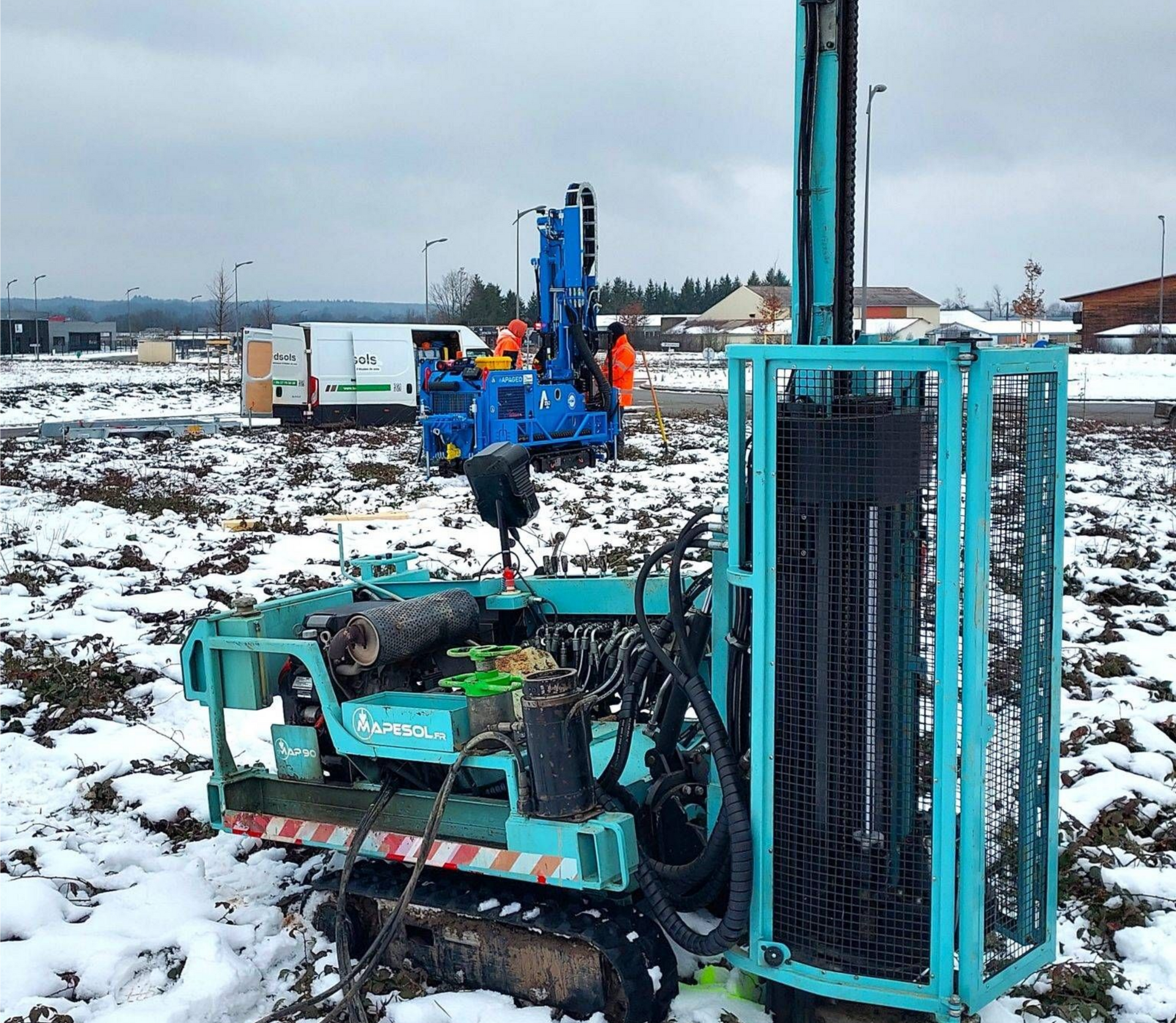 Équipement de forage bleu et vert sur un sol enneigé avec un chantier de construction en arrière-plan.