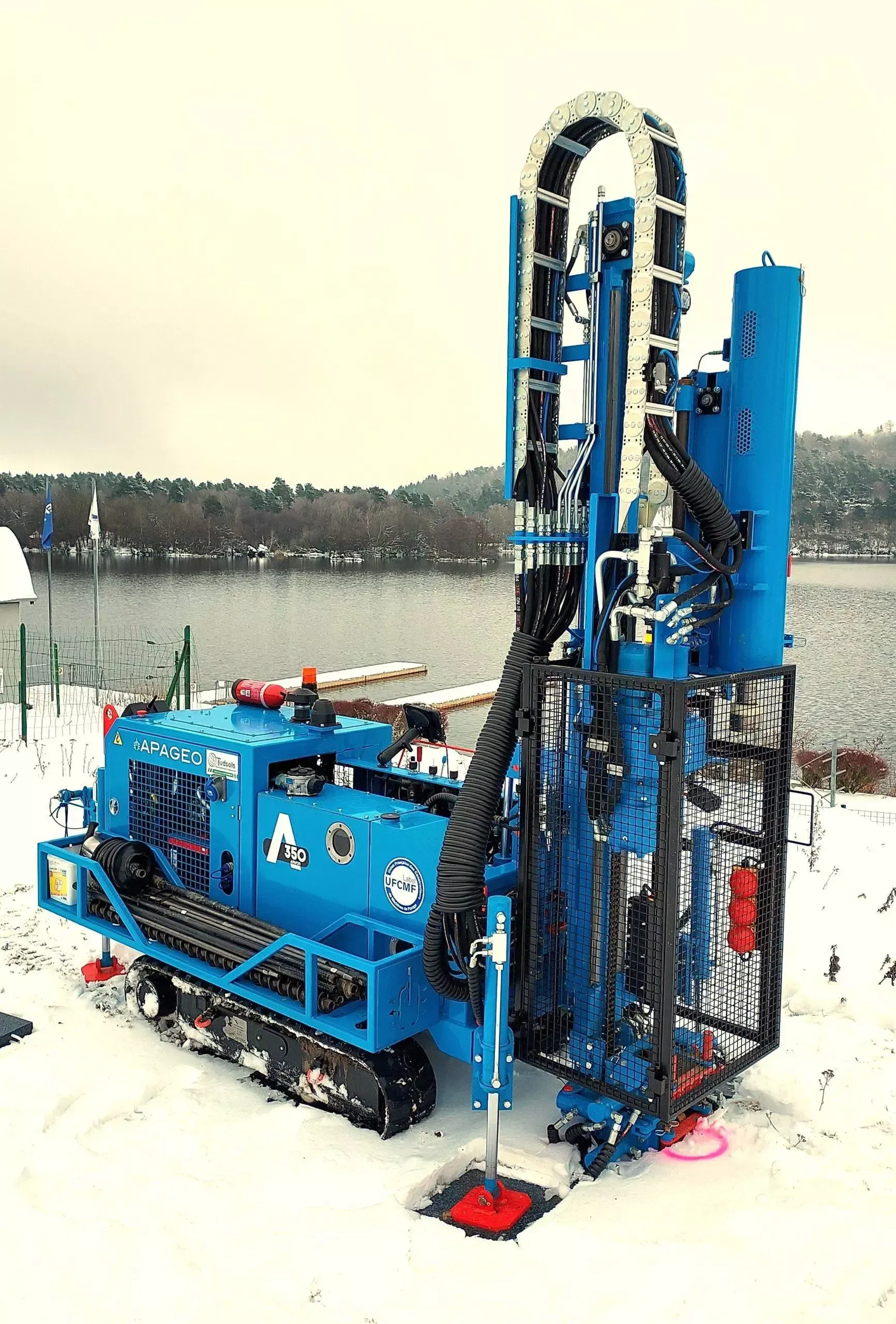 Foreuse bleue sur chenilles, avec un engin surélevé, travaillant sur un sol enneigé près de l'eau.
