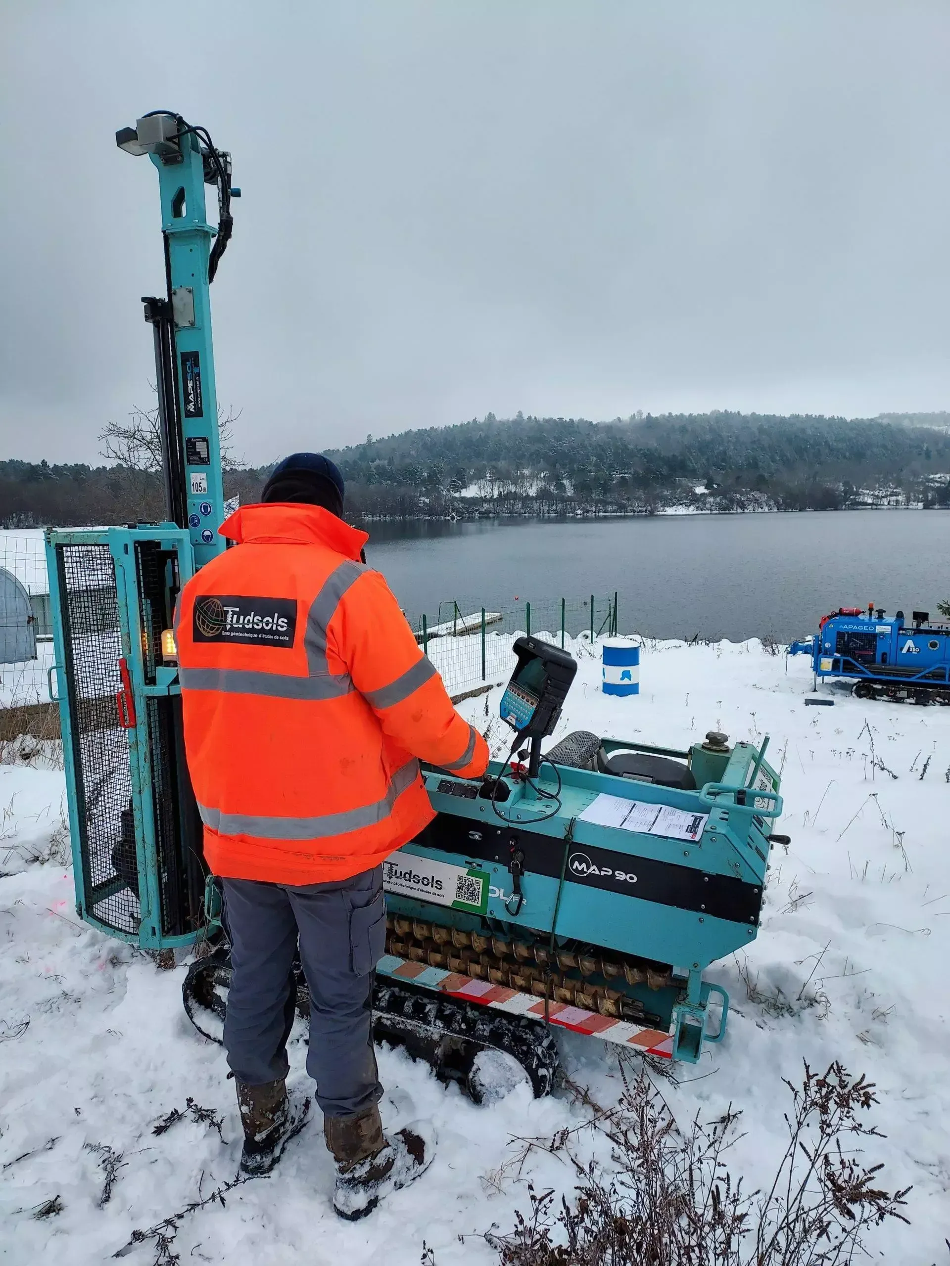 Une personne portant une veste orange utilise une foreuse bleu sarcelle sur un sol enneigé près d'un lac.