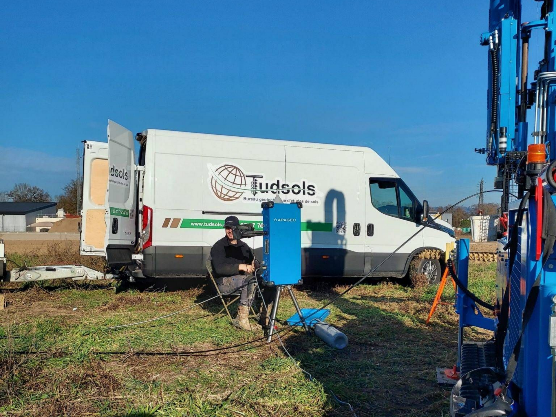 Une camionnette blanche avec le logo de l'entreprise garée dans un champ avec une personne est assise près d'une machine bleue.