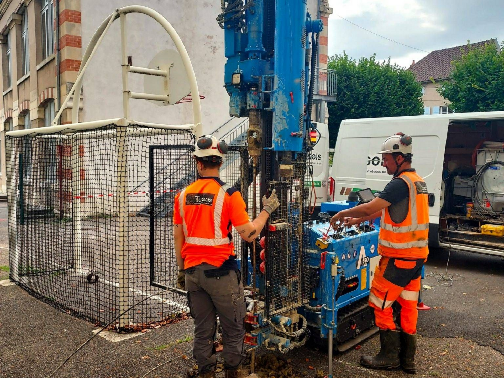 Deux ouvriers utilisant une perceuse bleue dans une rue de la ville avec les gilets et casques de sécurité orange.