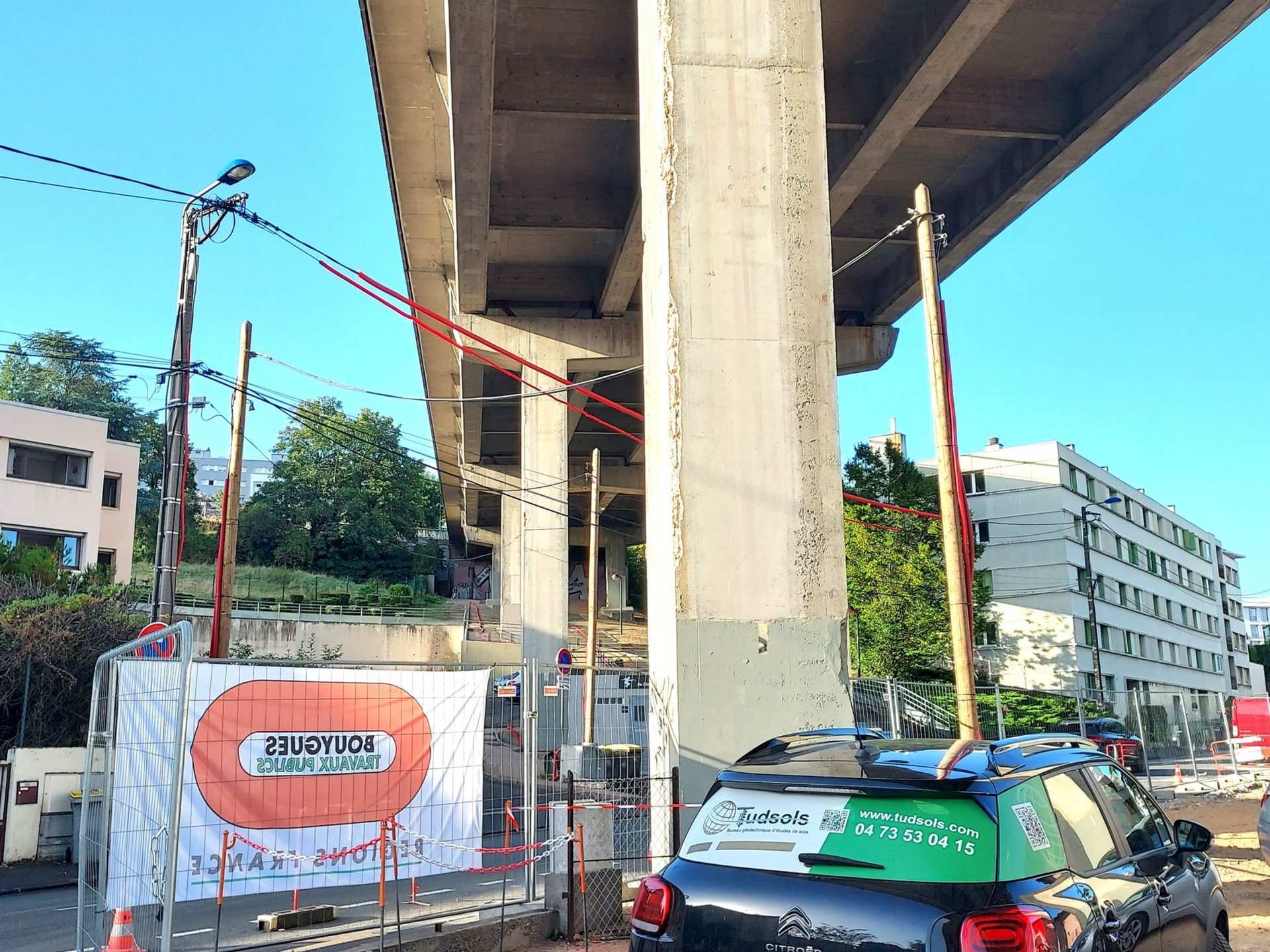 Chantier de construction sous un pont autoroutier avec une voiture et une clôture temporaire.