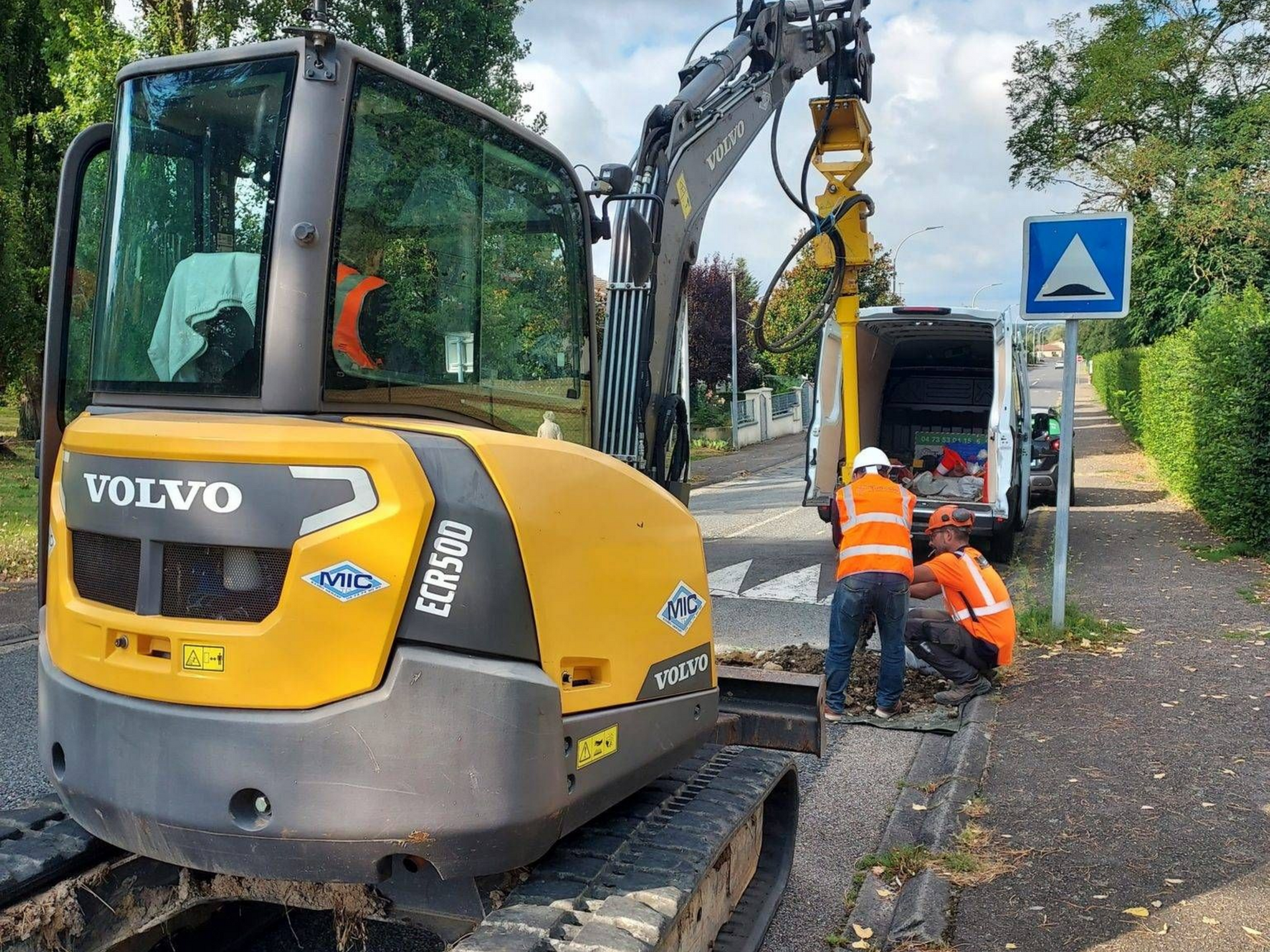Excavatrice Volvo jaune avec des ouvriers en gilet orange, travail dans la rue.