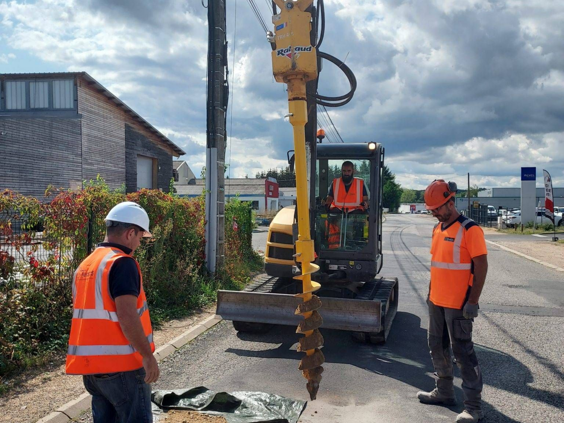 Des ouvriers du bâtiment forent un trou à l'arrière dans une rue et deux ouvriers portent des gilets orange et des casques de chantier.