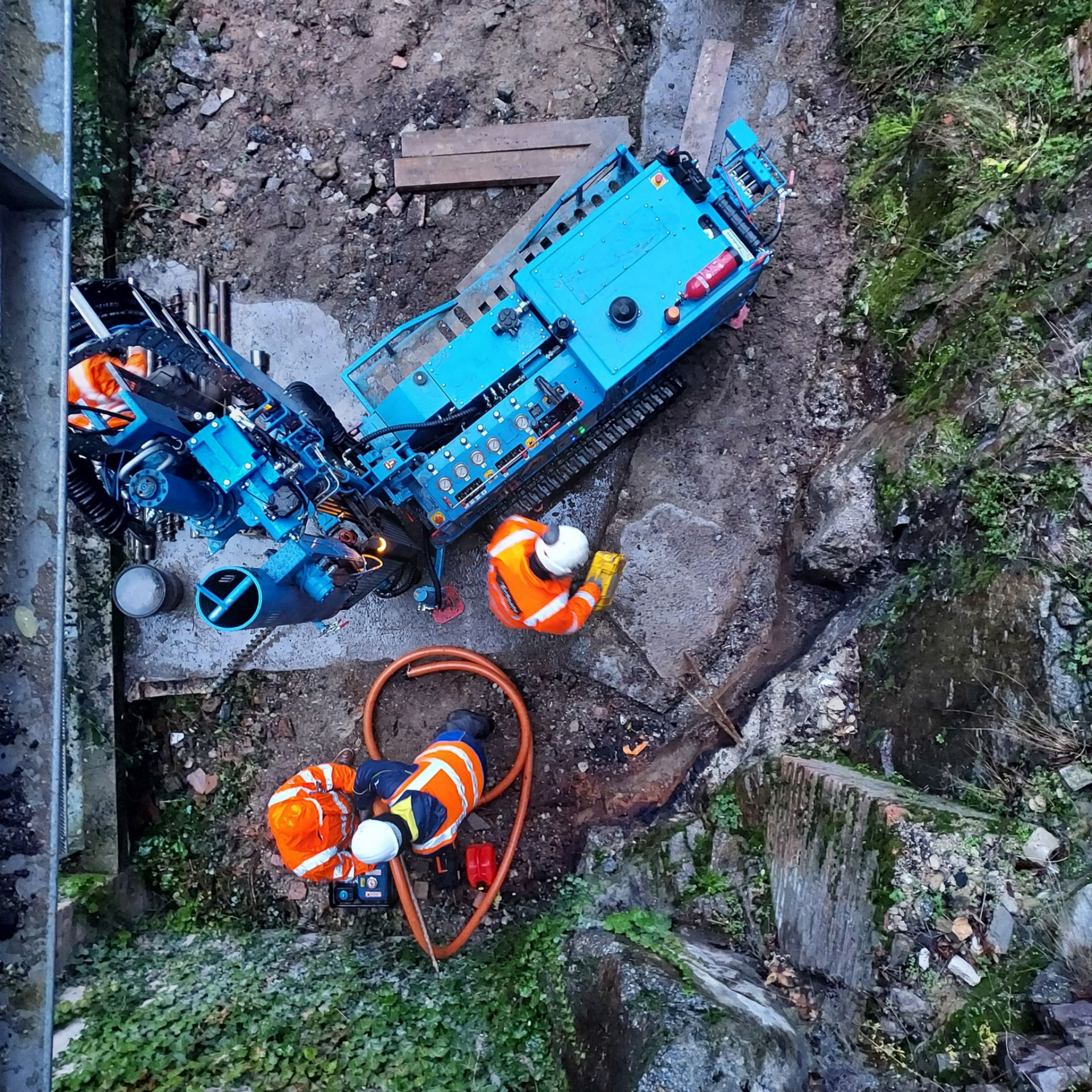 Vue aérienne : Des ouvriers du bâtiment en gilets orange manœuvrent une foreuse bleue dans une zone de terre avec de la végétation.
