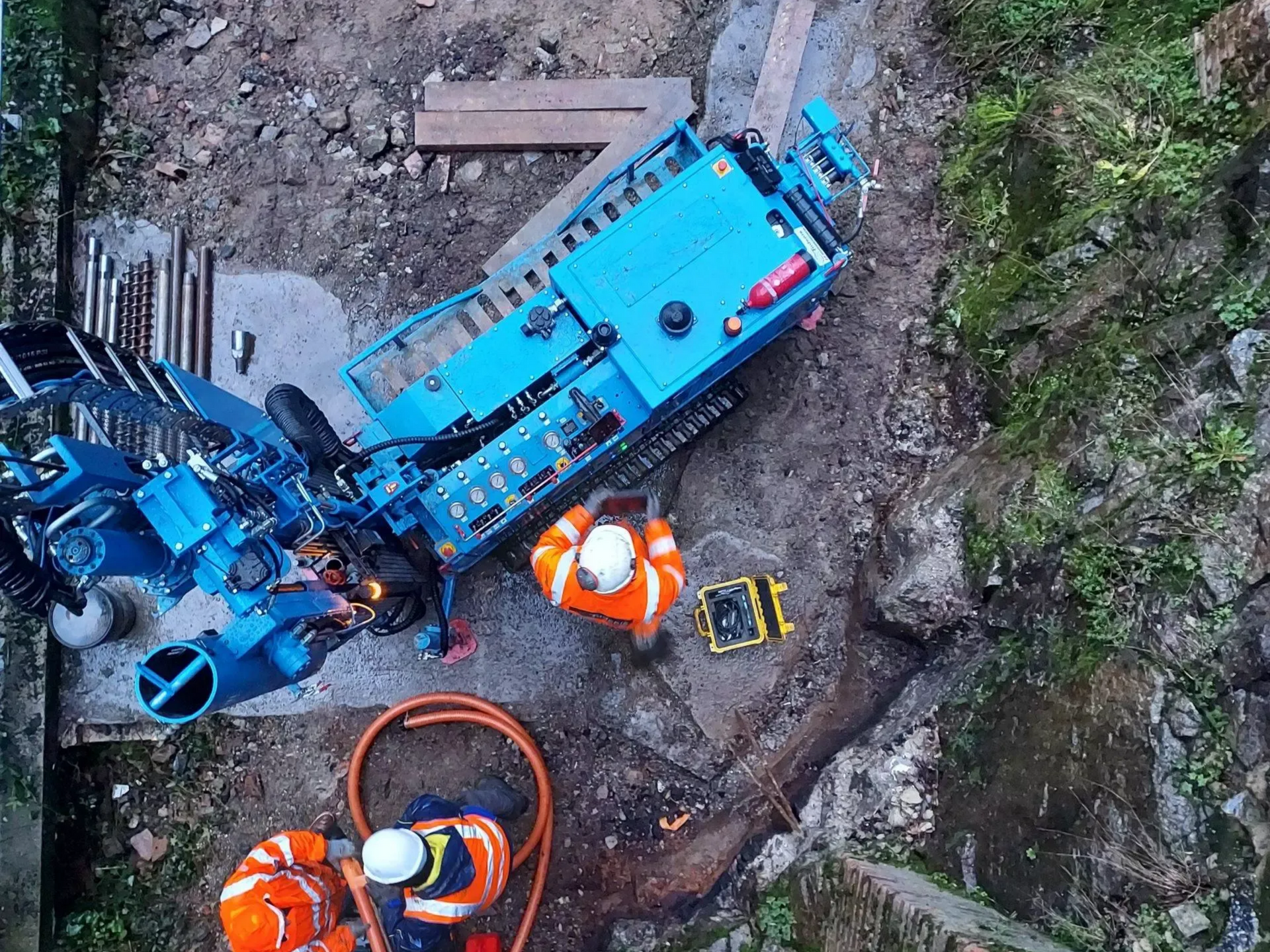 Vue aérienne d'une perceuse bleue et de deux ouvriers portant des gilets orange sur un chantier de construction.