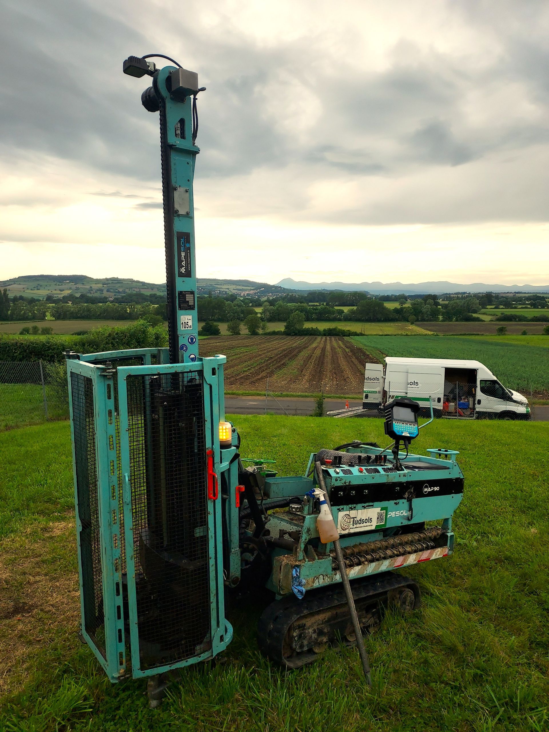 Foreuse bleue et turquoise sur chenilles dans un champ, haute tour de forage avec cage, ciel nuageux, véhicule en arrière-plan.