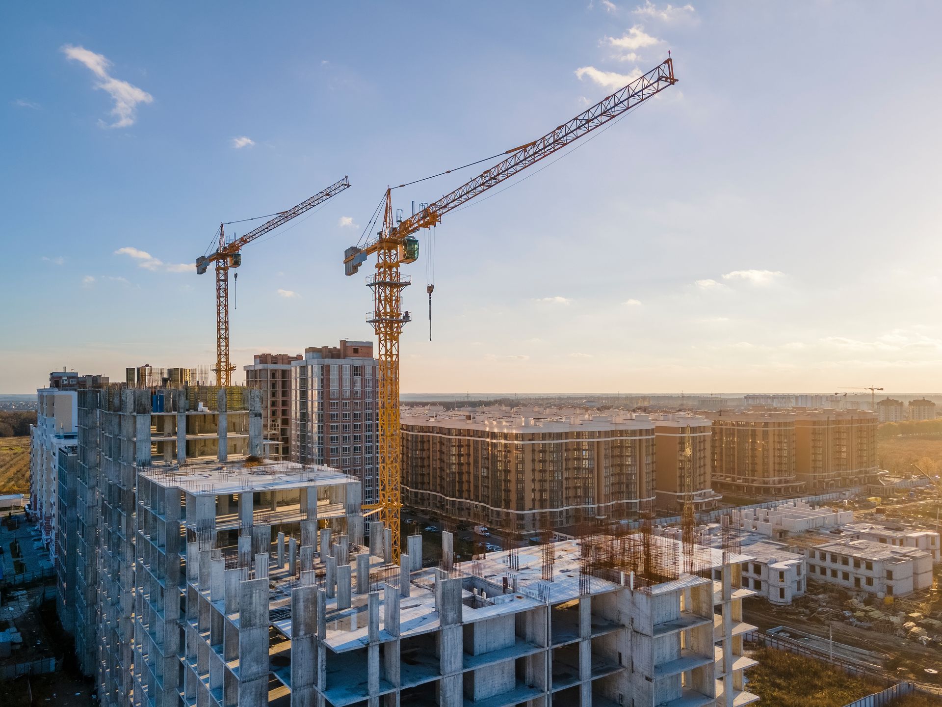 Chantier de construction avec grues, immeubles de grande hauteur partiellement construits, ciel bleu et autres bâtiments au loin.