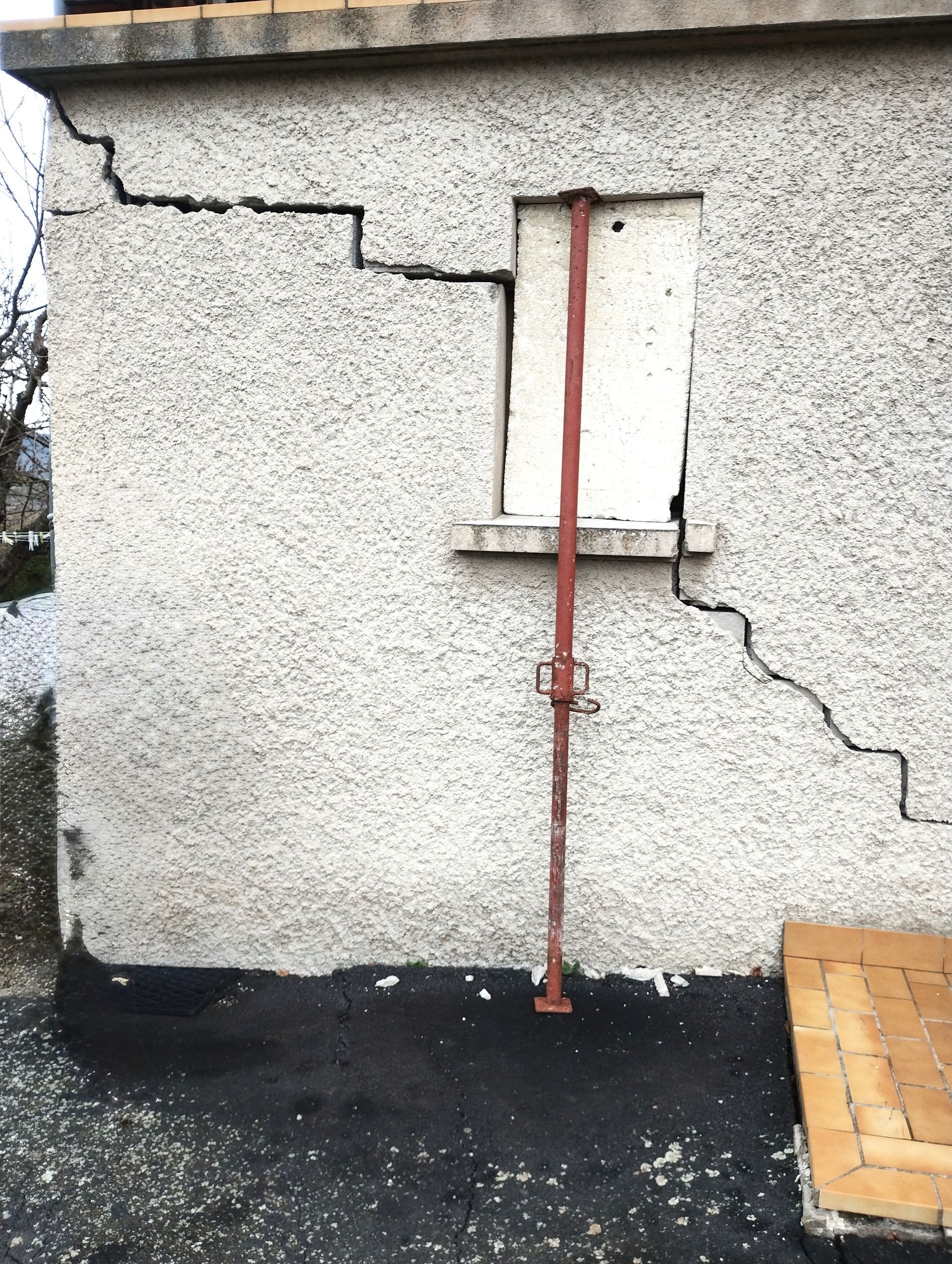 Mur extérieur présentant de larges fissures, fenêtre fermée et poutre de soutien métallique.