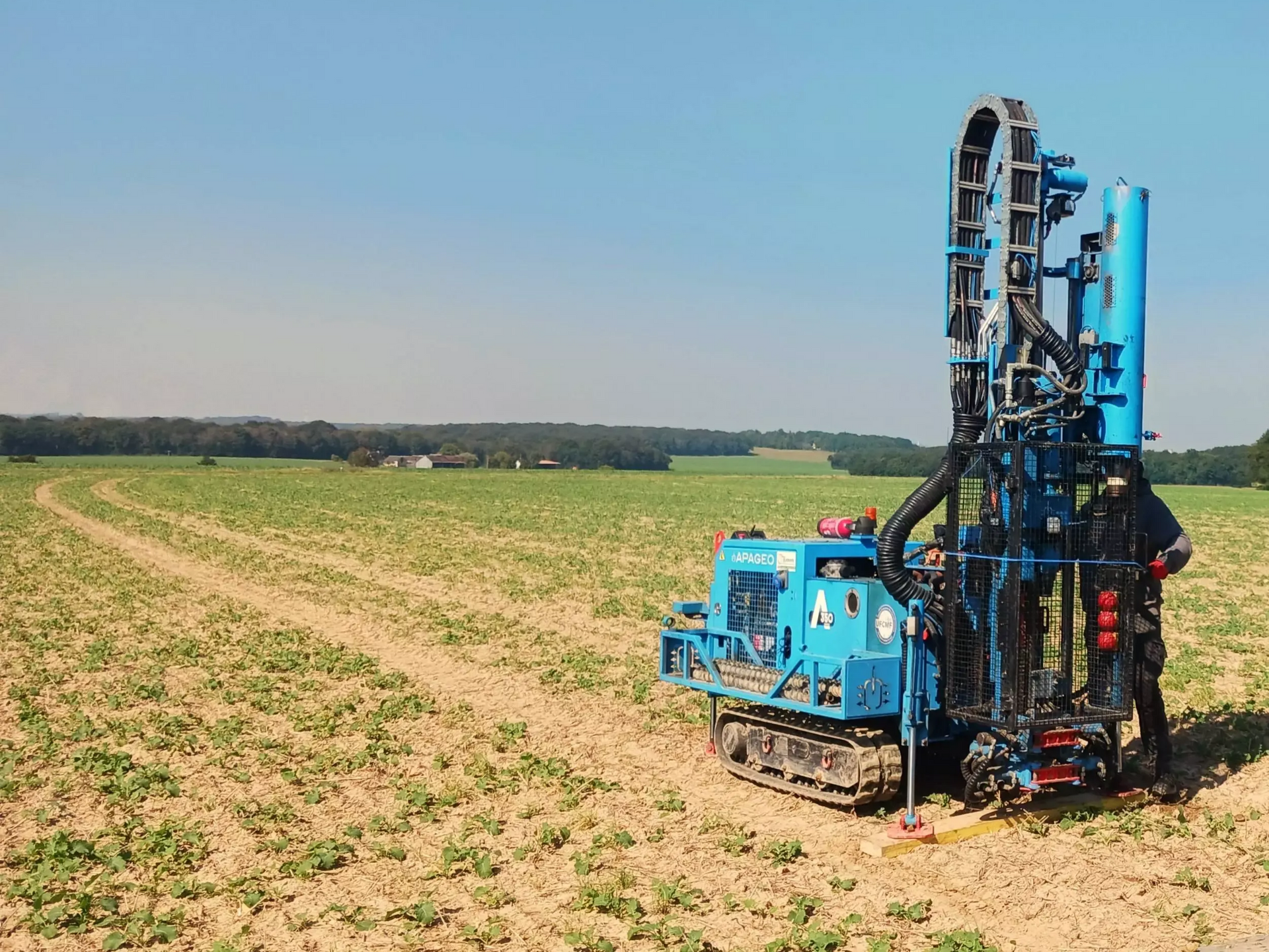 Foreuse bleue dans un champ, deux ouvriers à proximité, par une journée ensoleillée.