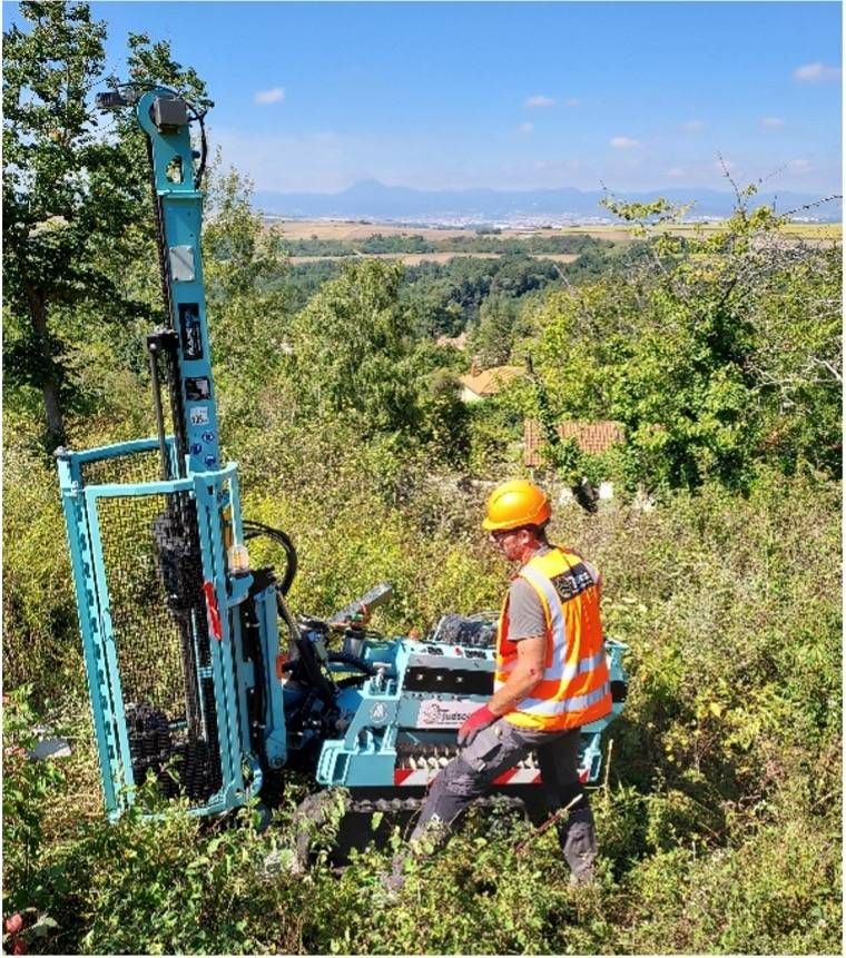 Un ouvrier en gilet orange opère une foreuse bleu sarcelle sur une colline, entourée de végétation verte.