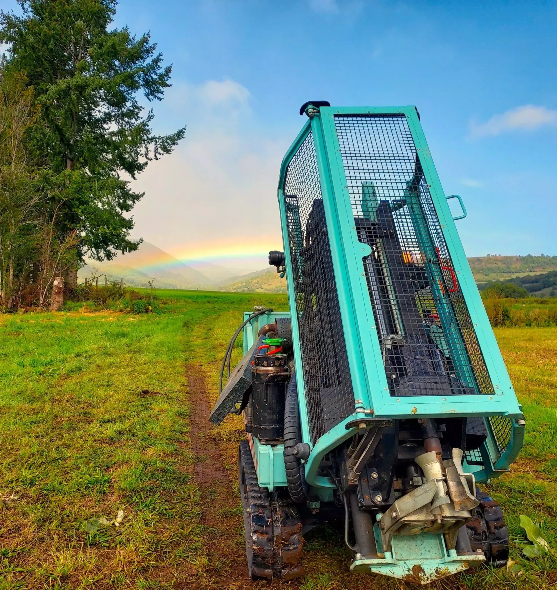 Véhicule à chenilles vert sur un champ herbeux avec un arc-en-ciel surplombant une zone boisée en arrière-plan.