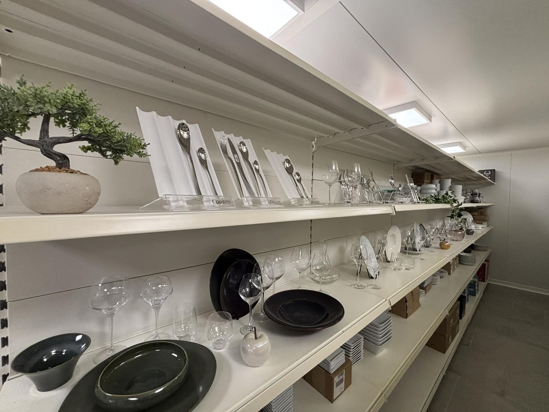 Des étagères présentent divers articles de vaisselle : assiettes, couverts et verres. Des tons blancs et gris dans un espace de rangement organisé.