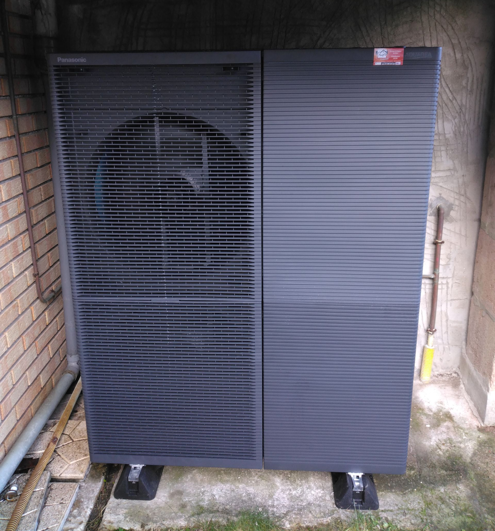 Unité de pompe à chaleur air-air gris foncé, à l'extérieur, montée sur des blocs noirs, contre un mur de briques.
