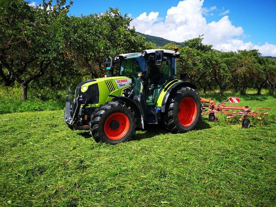 Un tracteur CLAAS vert vif et noir travaille dans un champ herbeux avec un râteau attelé, sur fond d'arbres.
