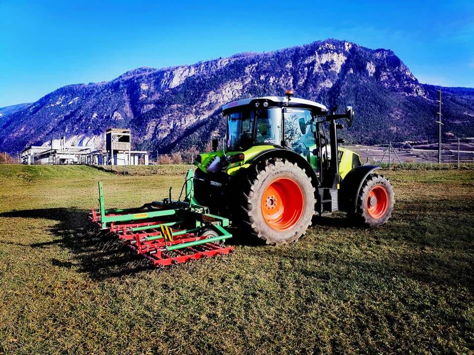 Tracteur vert à roues orange dans un champ, tirant du matériel agricole, avec des montagnes en arrière-plan.