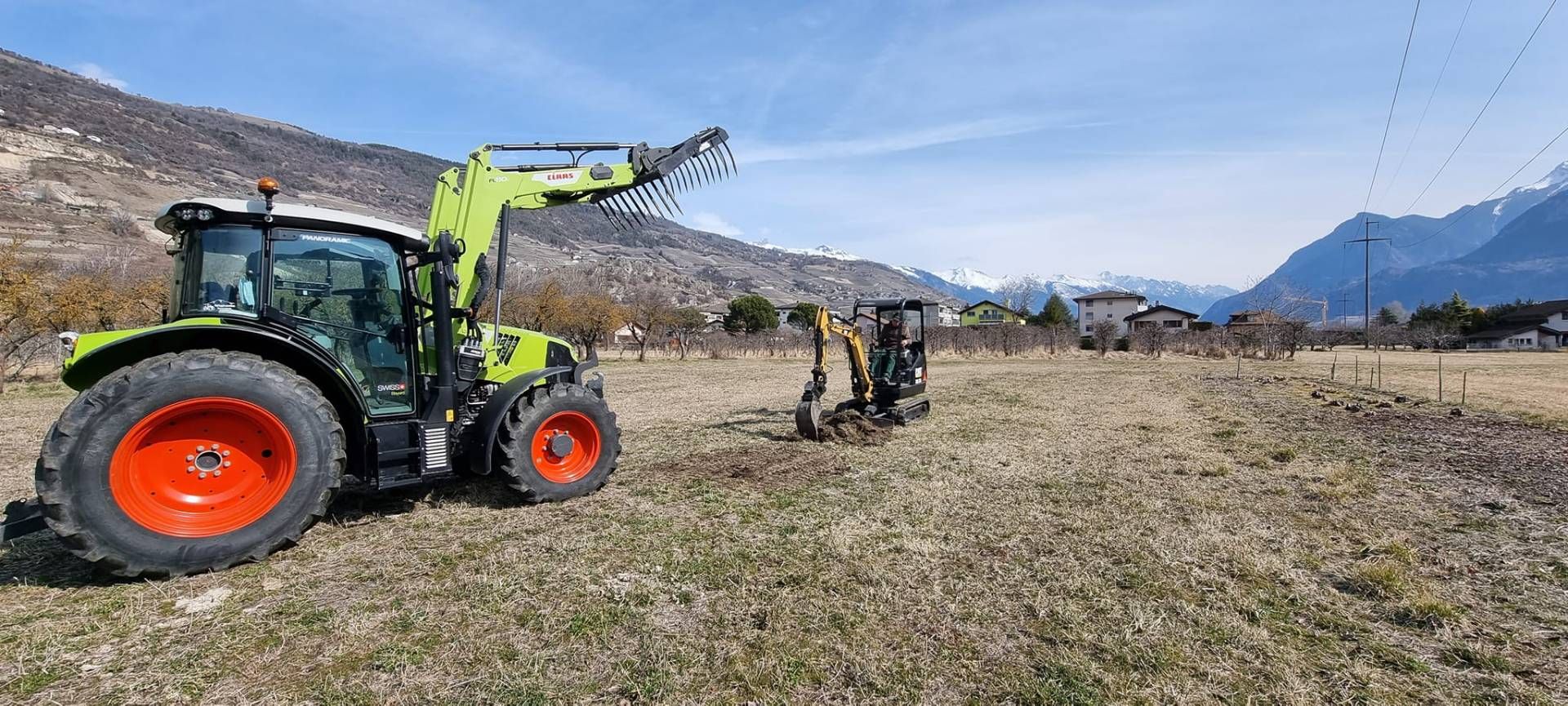 Un tracteur vert équipé d'un chargeur frontal et une petite pelle mécanique jaune travaillent dans un champ avec des montagnes en arrière-plan.