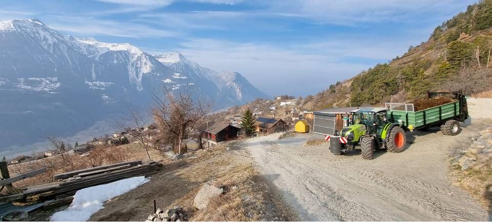 Un tracteur vert tirant une remorque est stationné sur un chemin de gravier dans un paysage montagneux aux sommets enneigés.
