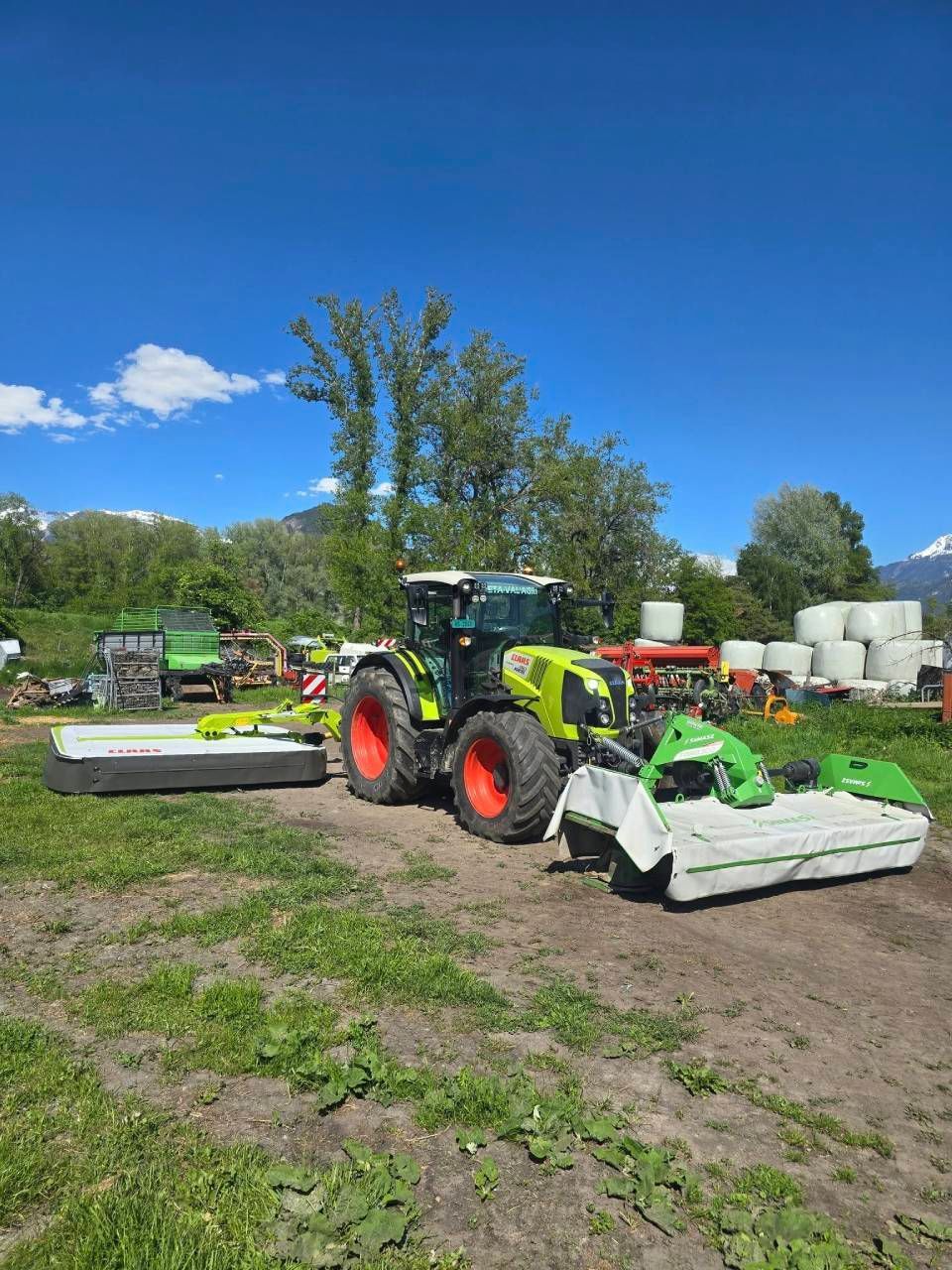 Un tracteur Claas vert et rouge vif, équipé de lames de fauche avant et arrière, est stationné sur un champ herbeux sous un ciel bleu.