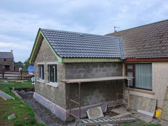Extension en parpaings partiellement construite avec toit de tuiles grises à côté d'une maison en construction.
