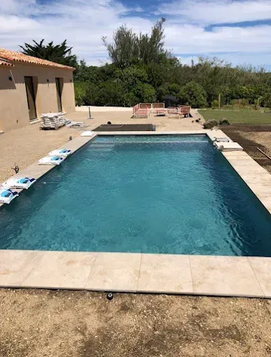 Piscine rectangulaire dans un jardin jouxtant une maison beige, avec mobilier de terrasse et arbres en arrière-plan.