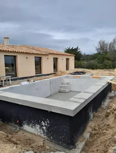 Piscine enterrée rectangulaire en construction à côté d'une maison beige, avec étanchéité noire et marches blanches.